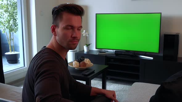 A Man Watches a TV with a Green Screen, Then Turns To the Camera and Shakes His Head, Unhappy