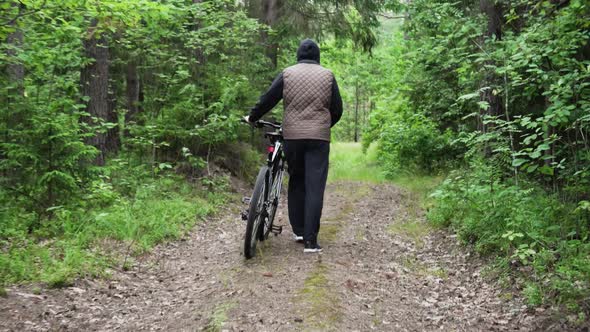 Man Walks and Rolls a Bicycle Next to Him Along a Forest Path