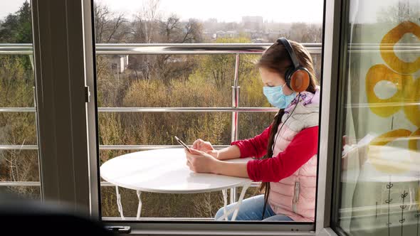 Teenager Girl in Mask and Headphones, Uses Smartphone, Makes Selfie, on Open Balcony. Spring Sunny