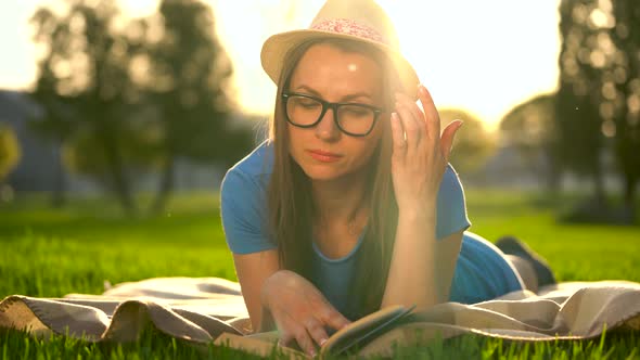 Girl in Glasses Reading Book Lying Down on a Blanket in the Park at Sunset