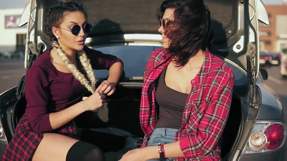 Two Young Attractive Women in Stylish Sunglasses Sitting in Car Trunk in the Parking By the Shopping