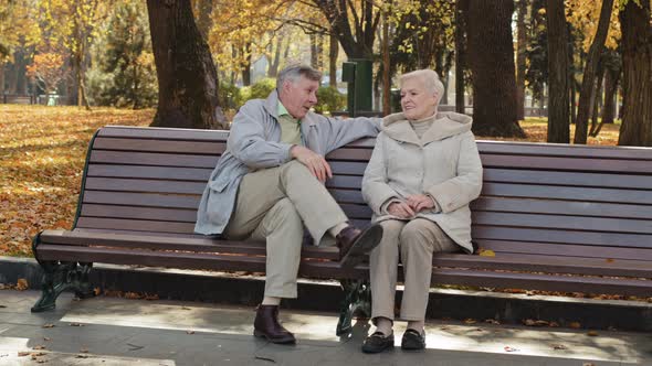 Mature Old Caucasian Couple Grandparents Sit on Bench Senior Pensioner Man Cuddling Closer to Middle
