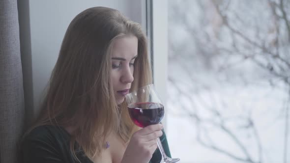 Close-up Portrait of Young Sad Caucasian Woman Drinking Wine and Looking Out the Window Indoors