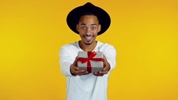 Excited Hipster African Man in Hat Holding Gift Box and Gives It By Hands To Camera on Yellow Wall