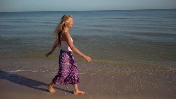 Pretty mature woman smiling and walking along a beach dressed in a sarong and looking very happy.