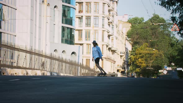 Young Man in Casual Clothes Glasses and Hat is Skateboarding Along Deserted City Street Surrounded