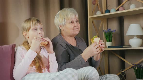 Active Elderly Woman with Granddaughter Together Watching TV and Eating Sandwiches