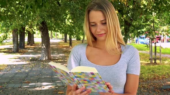 Young Pretty Blond Woman Reads Book - Park with Trees in Background