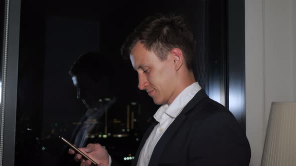 Young Businessman in Office Suit Is Browsing Smartphone Near the Window with City View.