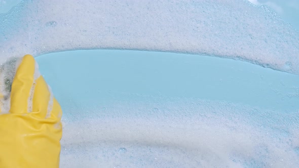 Hand in a Yellow Rubber Glove Holds a Cleaning Sponge and Wipes a Soapy Foam on a Blue Background