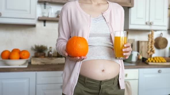 Closeup Of Pregnant Woman Belly With Glass Of Freshly Squeezed Orange Juice In Hand In Kitchen