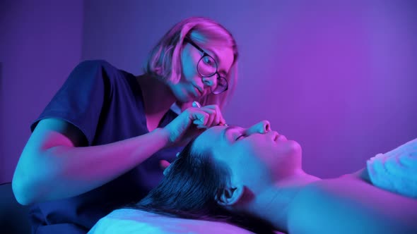 Massage Session  Young Woman Using Jade Roller on the Face of Her Female Client in Purple Lighting