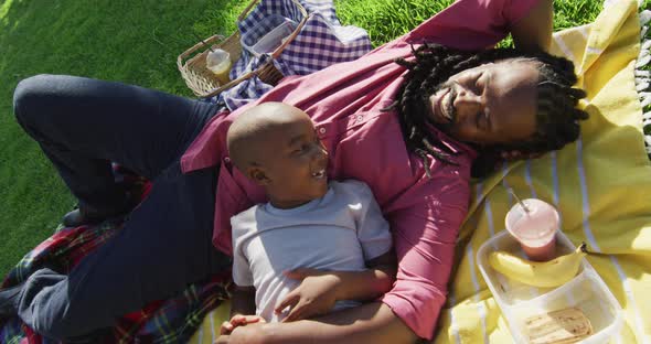 Video of happy african american father and son having picnic on grass and resting on blanket