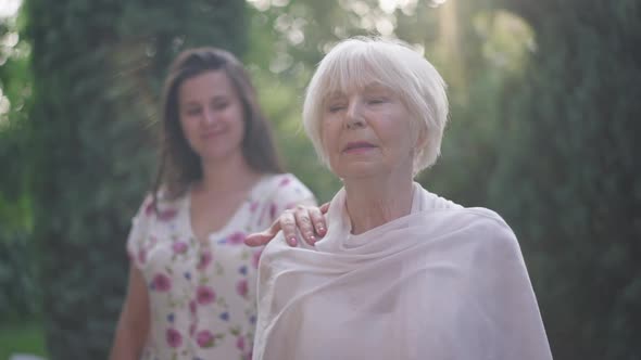 Beautiful Senior Elegant Woman Standing in Sunrays in Spring Summer Park with Blurred Granddaughter