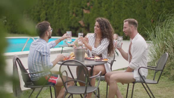 Group of happy young people cheering with drinks and eating fruits by the pool in the garden