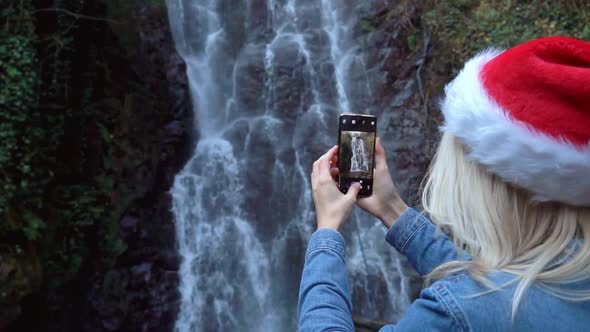 blonde in Santa Claus hat stands waterfall takes pictures photos on smartphone