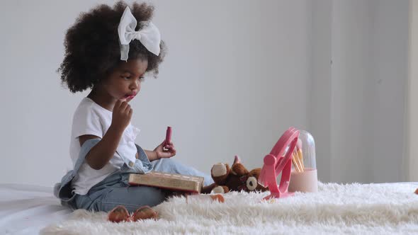 African preschool girl sitting on bed, having fun, and applying lip gloss