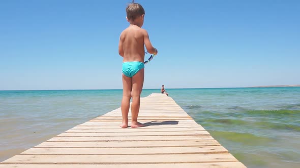 Small Tanned Boy with Light Hair, Dressed in Swimming Shorts Stands on a Wooden Pier on a Summer Day