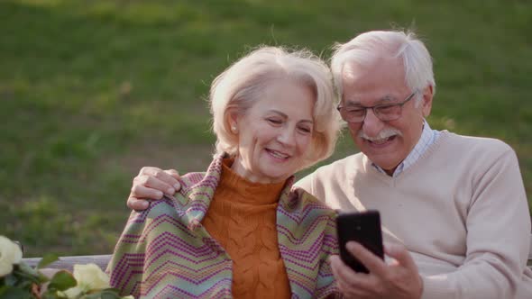 Handsome senior couple sitting on the bench with basket full of flowers and looking at mobile phone