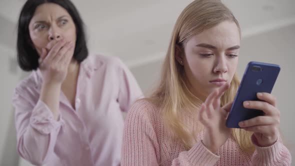 Portrait of Caucasian Mother Spying Teen Daughter. Blond Girlhiding Smartphone and Turning Back