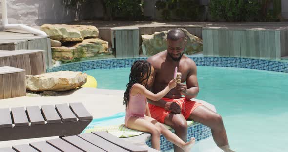 Happy african american father and daughter eating ice creams at swimming pool