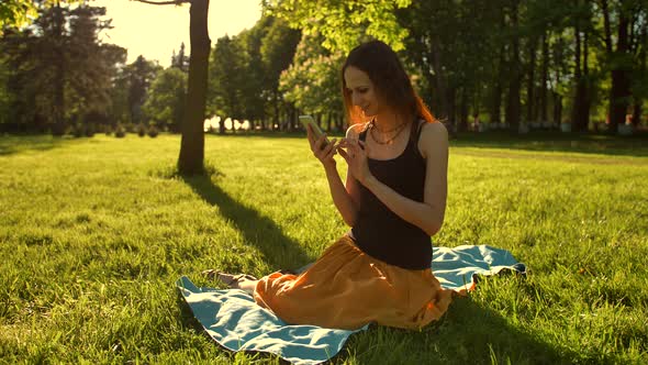 Sexy Red Hair Girl Enjoying Sunny Day. Sitting on Bench in Green Summer Park Using Mobile.