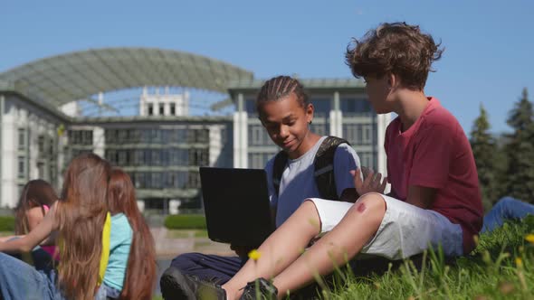 Multiethnic Schoolboys Study Together on Laptop Sitting on Grass in Park