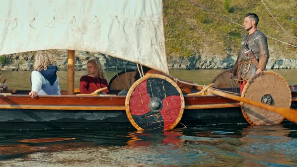 Vikings Sail on an Old Ship with a Raised Sail Along a Calm River