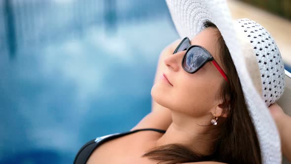 Pretty Woman in Sunglasses and Hat Enjoying Sunbathing Lying on Deck Chair Near Swimming Pool