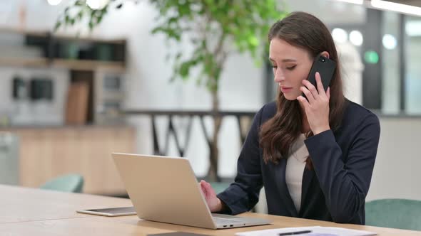 Businesswoman with Laptop Talking on Smartphone at Work