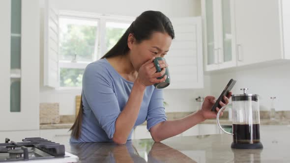 Happy asian woman standing at table, drinking coffee and using smartphone in kitchen