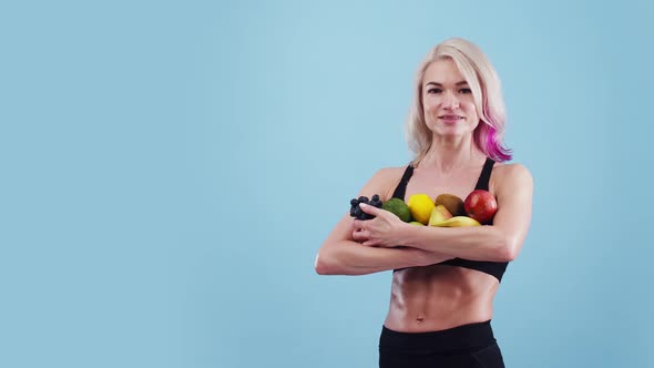 Woman of athletic physique holds a pile of fruit in hands and smiles in the studio, blue background.