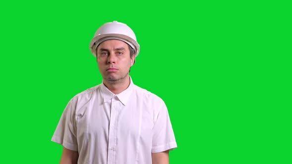 Young Male Angry Engineer in White Hardhat and Shirt Standing and Knocking the Helmet on Green