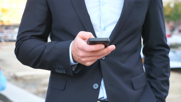 Young Successful Businessman in Suit Standing Among City Street and Browsing Smartphone. Confident