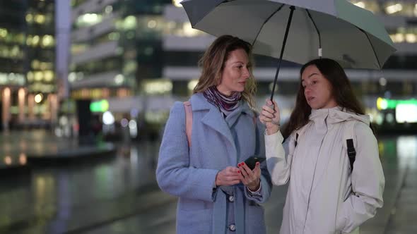Two Women Stand Under an Umbrella and Chat Against the Background of a Blurred Evening Street and
