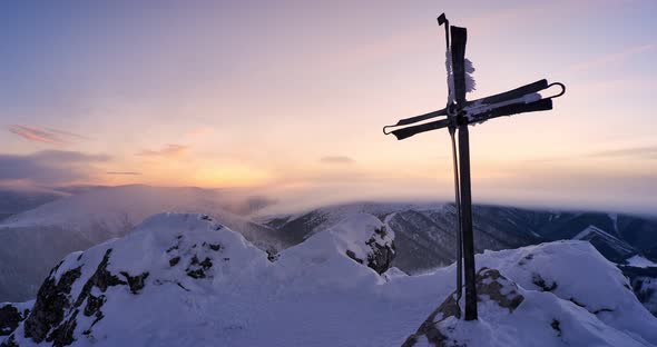 Christian cross in a snowy mountain landscape. Clouds spill over the hills at dusk.