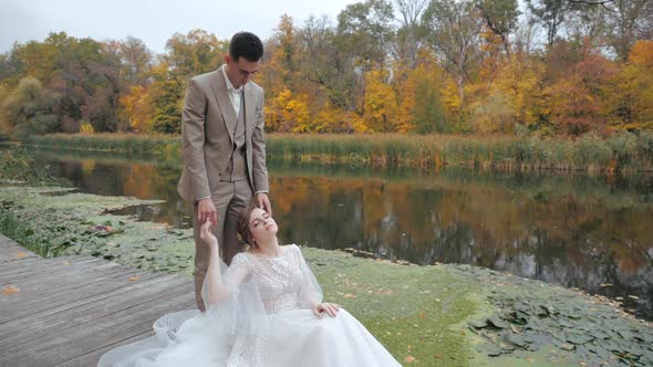 Beautiful Young Bride in a Wedding Dress Is Sitting on Wooden Bridge with Eyes Closed, Leaning on