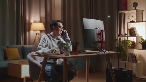 Asian Male Footwear Designer Talking On Smartphone While Working On A Desktop Computer