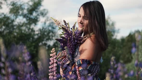 Young Beautiful Smiling Woman with a Bouquet Walks Among the Flowers of Purple Lupins