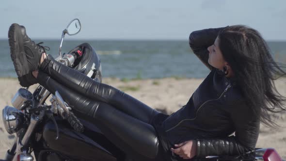 Pretty Young Caucasian Woman in a Black Leather Jacket and Pants Lying on a Motorcycle Near Sea