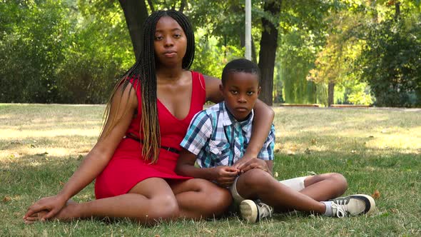 A Young Black Mother and Her Son Sit on Grass in a Park and Look Seriously at the Camera
