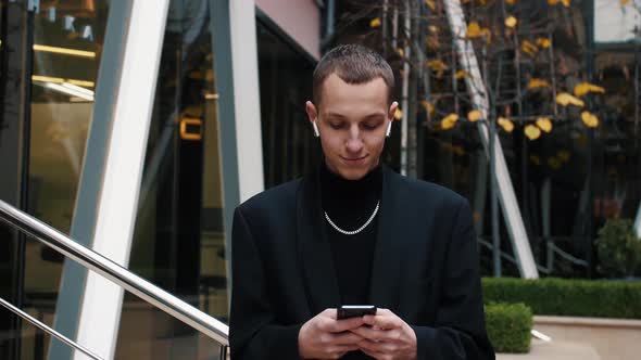 Young Businessman Using His Smartphone and Smiles. He Is Texting Messages While Standing Near Modern