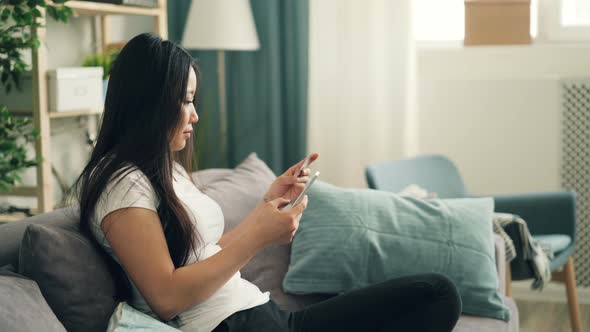 Attractive Asian Lady Is Using Smartphone To Make Online Payment with Bank Card Sitting on Sofa at