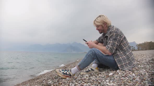 A Young Man in a Plaid Shirt Sits on the Shore and Uses His Phone