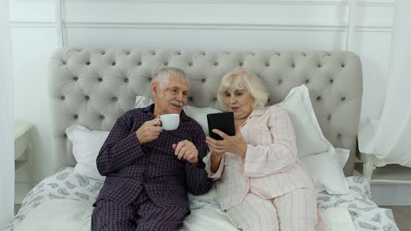 Senior Elderly Couple Wearing Pyjamas Lying on Bed Looking on Digital Tablet Laughing and Having Fun