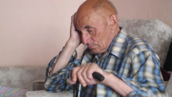 pensive caucasian pensioner 70-79 years old with a walking stick sits at home with his hand holding 