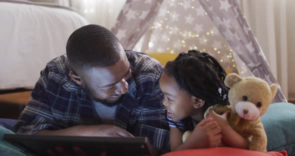 Happy african american father and daughter lying in tent and using tablet with copy space