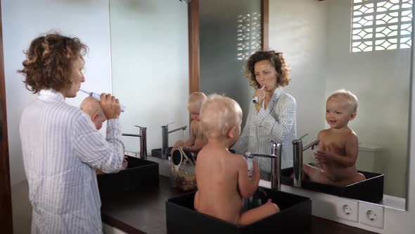 Young Mother Brushes Her Teeth with Her Baby Twins Sitting in the Sinks