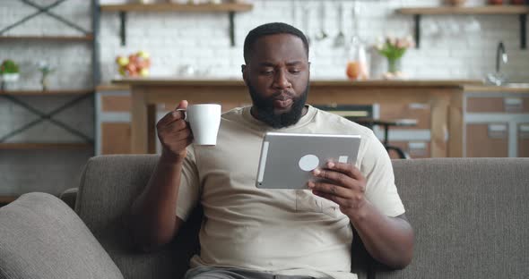 African Man Watching Video on Tablet PC and Drinking Coffee While Sitting on Sofa at Home