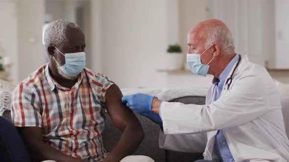 Senior caucasian male doctor vaccinating senior african american man at home both wearing face masks